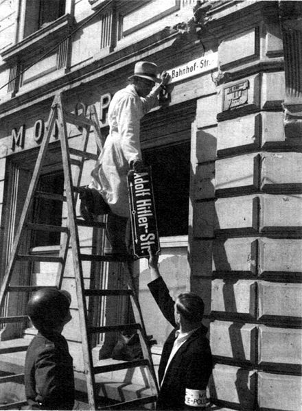 Tausch der Straßenschilder in Trier im Mai 1945. Die zur Zeit des Nationalsozialismus in "Adolf-Hilter-Straße" umbenannte "Bahnhofstraße" erhält ihren alten Namen wieder.